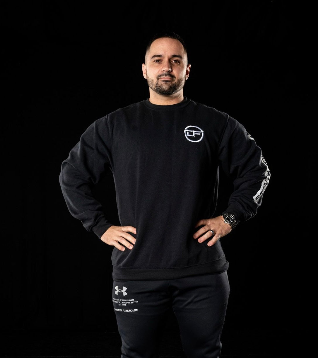 Image of Josh DeMelo, owner and head trainer of DeMelo Fitness. Josh is standing with his arms at his hips against a black background wearing a DeMelo Fitness branded long-sleeved shirt