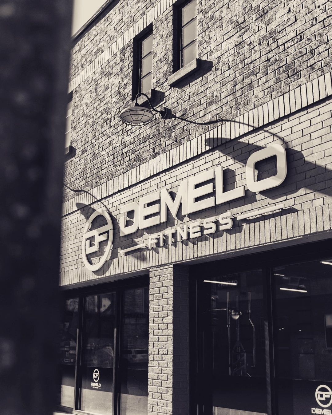Exterior shot of the DeMelo Fitness gym. The photo is sepia toned showing the logo and brand name along the wall and in the corner of the windows