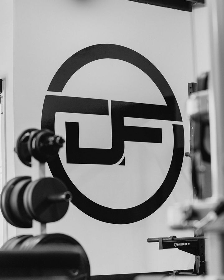 A large DeMelo Fitness logo displkayed along a white wall with weights in the foreground blurred out
