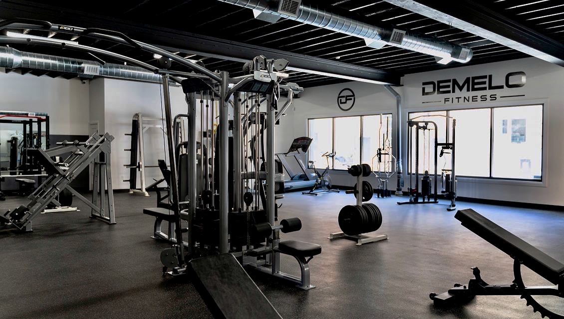Wide shot of the DeMelo Fitness facility. Along the walls are branding of the gym, displaying logos and the name of the location: DeMelo Fitness. In the foreground are multiple pieces of gym equipment, including benches, leg lift machinery, and weights. In the background are cardio machines like a treadmill.