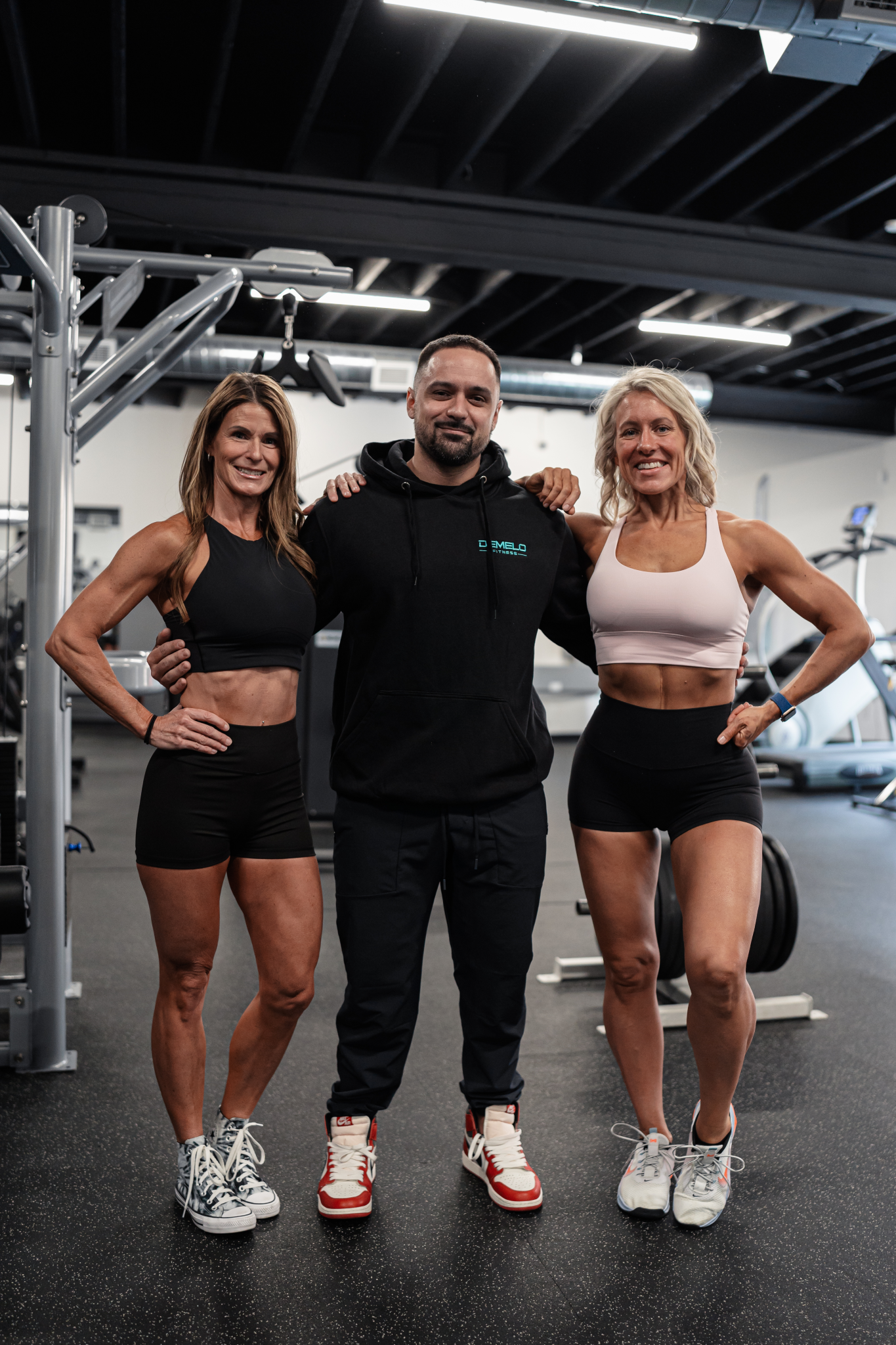 Three confident builders standing side by side. In the center is Josh DeMelo, owner and head trainer of DeMelo Fitness. Josh has two women beside him, one on either side.
