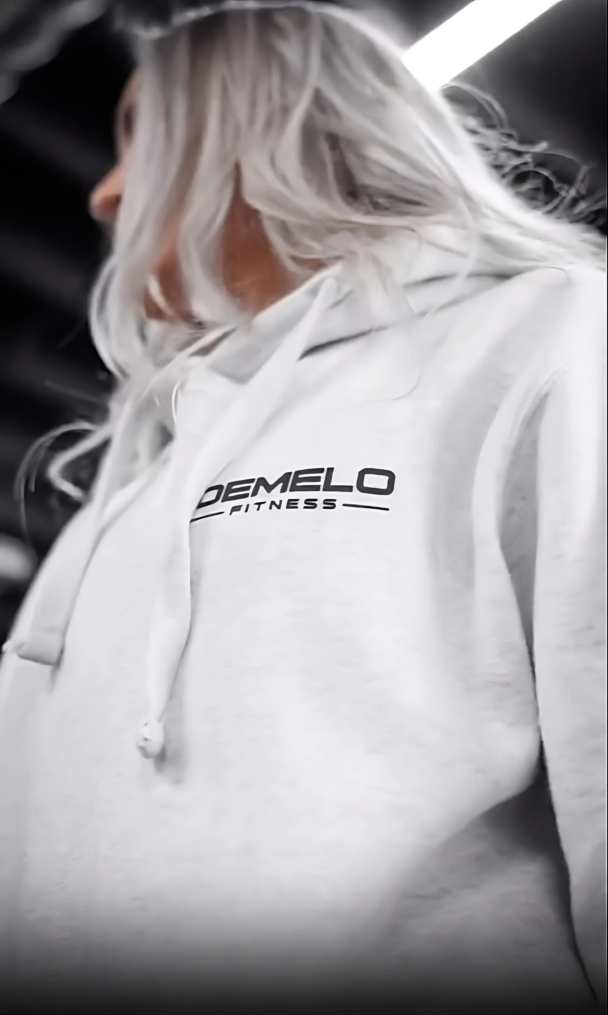 A photo of a woman with white hair wearing a white DeMelo Fitness hoodie