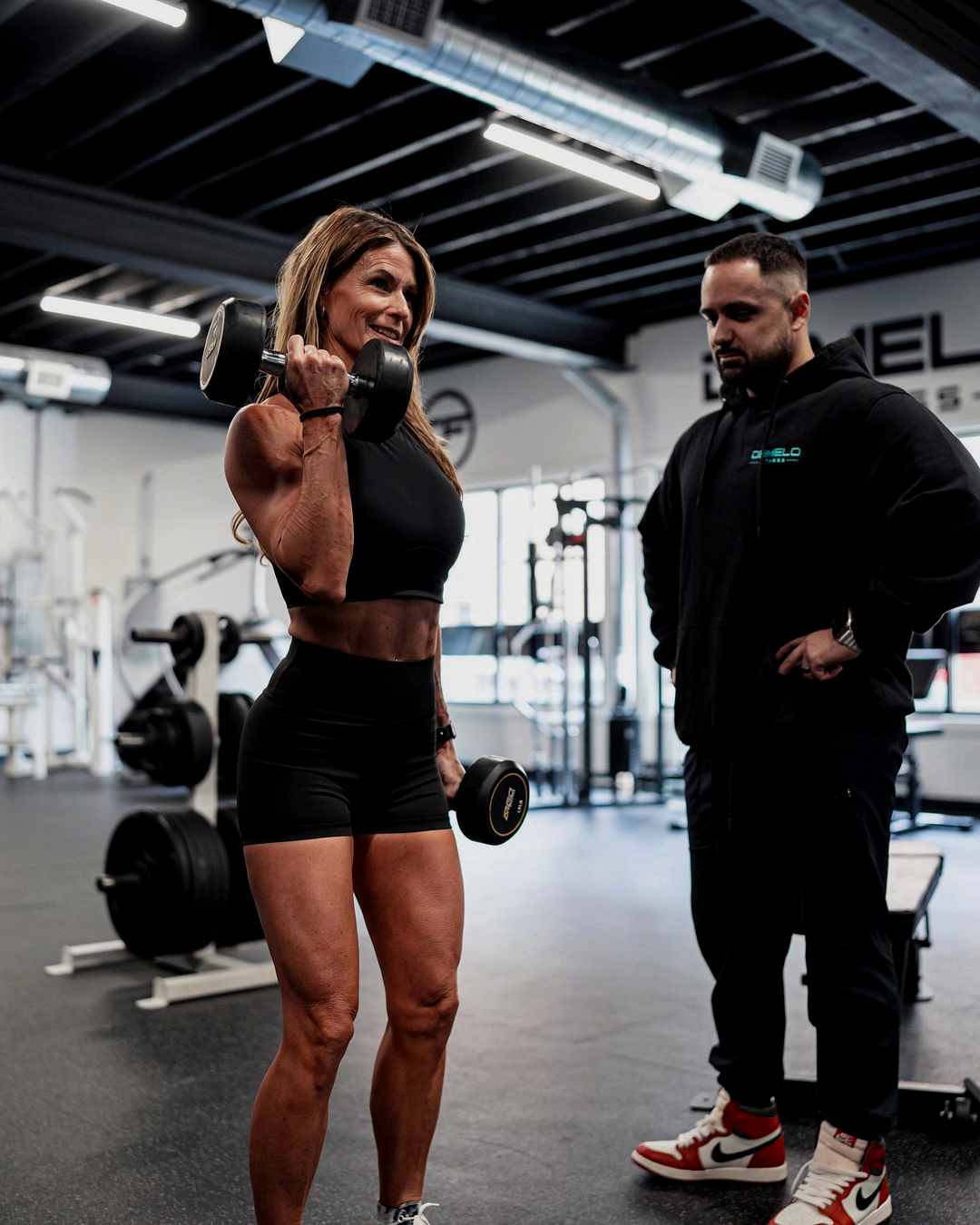 A woman is lifting hand weights in either hand while Josh DeMelo spots her, making sure form is correct and progress is being made